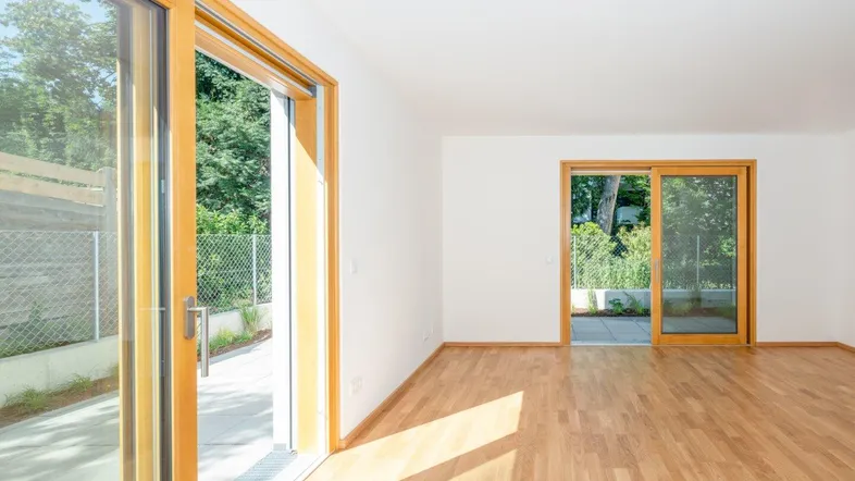 Helles Zimmer mit Holzboden und großen Glasschiebetüren, die zu einer Terrasse und Grünfläche führen.