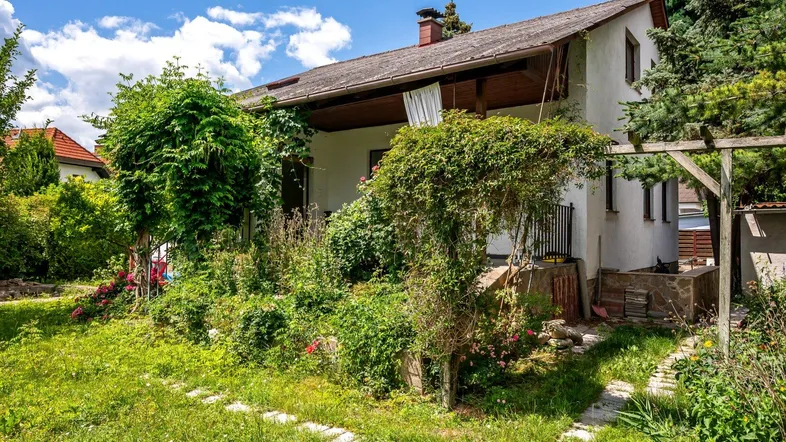 Charmantes Haus mit üppigem Garten und überdachter Terrasse unter blauem Himmel.