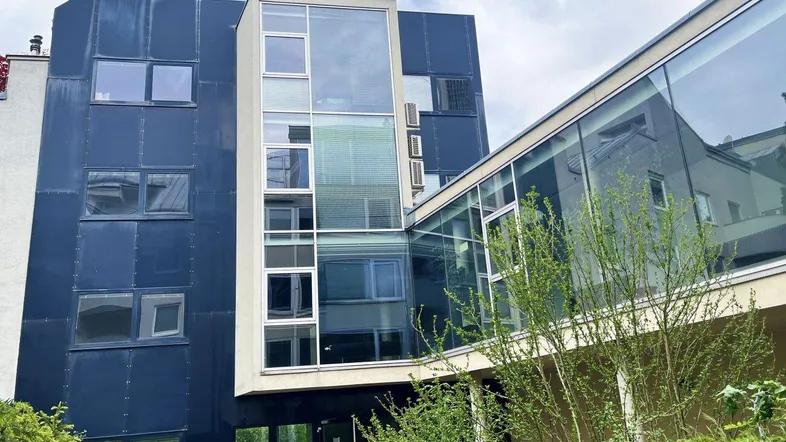 Moderne Gebäudefassade mit dunklen Paneelen und großen Glasflächen, umgeben von Grünpflanzen.