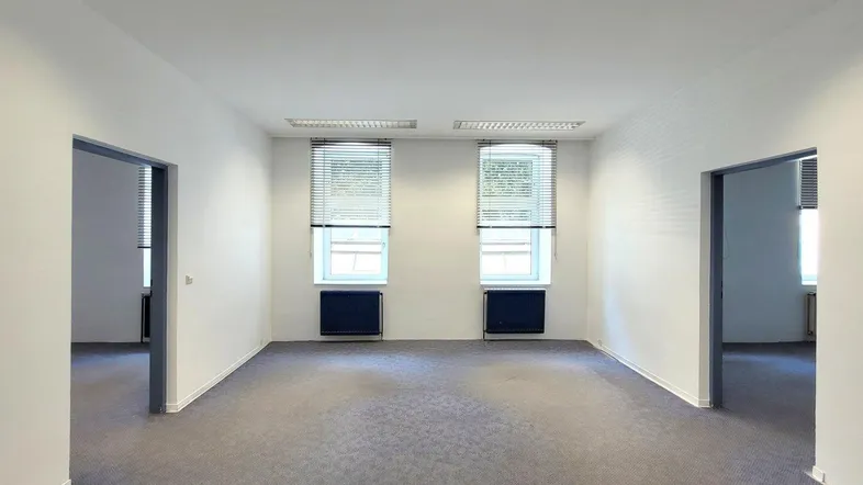Geräumiges, leeres Büro mit grauem Teppichboden, weißen Wänden und zwei Fenstern mit Jalousien.