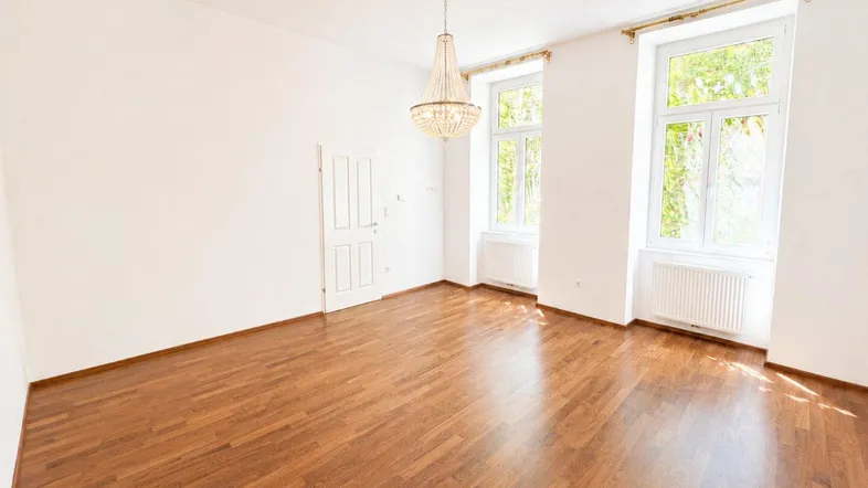 Heller, leerer Raum mit elegantem Kronleuchter, Holzboden und hohen Fenstern.