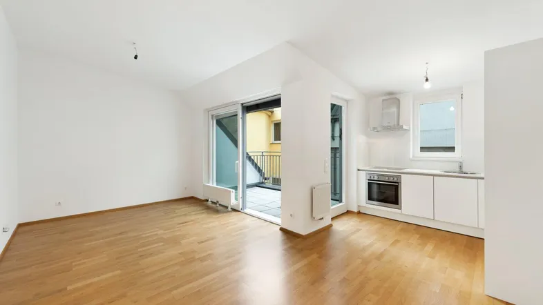 Heller offener Wohnbereich mit Parkettboden, integrierter Küche und Zugang zum Balkon.