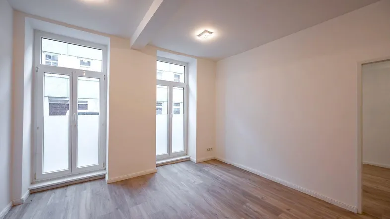 Leeres Zimmer mit hellem Laminatboden und zwei großen Fenstern, bereit zur individuellen Gestaltung.
