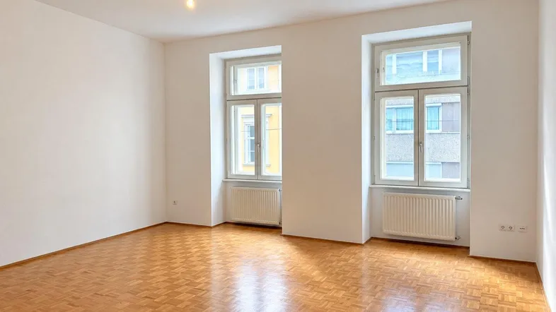 Helles Zimmer mit Parkettboden und zwei großen Fenstern, die viel Tageslicht hereinlassen.