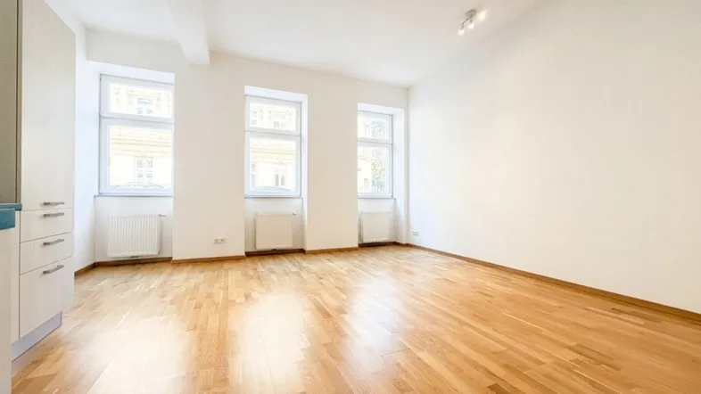 Helles, leeres Zimmer mit Holzboden und drei Fenstern, bereit zur individuellen Gestaltung.