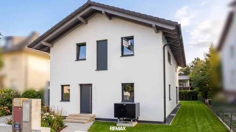 Modernes Einfamilienhaus mit weißer Fassade, dunklem Dach und Wärmepumpe im Vorgarten.