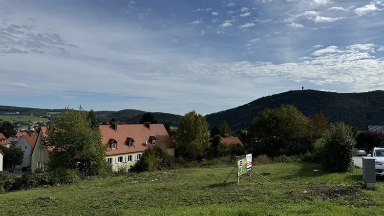 Weitläufige Grünfläche mit Blick auf bewaldete Hügel und eine ländliche Siedlung unter blauem Himmel.
