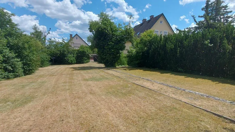Große Gartenfläche mit trockenem Rasen und umgebenden Bäumen, im Hintergrund sind Häuser sichtbar.