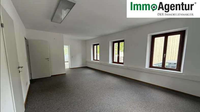 Geräumiger Büroraum mit grauem Teppichboden, weißen Wänden und mehreren Fenstern für Tageslicht.