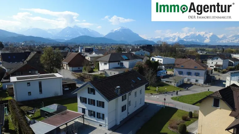 Luftaufnahme eines Wohnhauses mit umliegenden Häusern und schneebedeckten Bergen im Hintergrund.