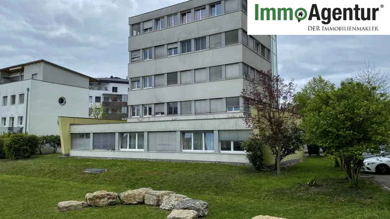 Mehrstöckiges Wohngebäude mit grauer Fassade und Grünfläche im Vordergrund unter bewölktem Himmel.