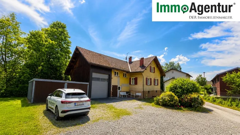 Traditionelles Einfamilienhaus mit gelber Fassade, rotem Dach und großem Kiesparkplatz unter blauem Himmel.