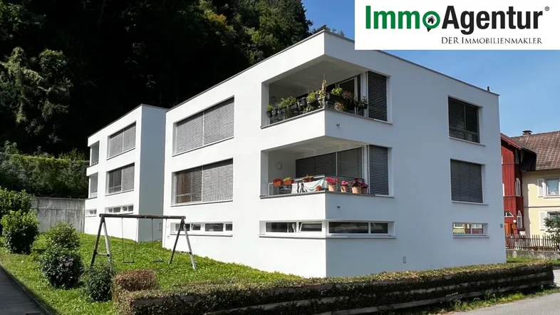 Modernes weißes Mehrfamilienhaus mit Balkonen und einem kleinen Spielbereich im Grünen.