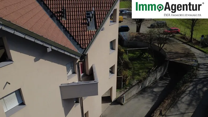 Luftaufnahme eines Mehrfamilienhauses mit Ziegeldach und kleinem Balkon, umgeben von Grünflächen und einer Zufahrt.