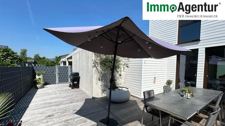 Geräumige Holzterrasse mit Esstisch, Sonnenschirm und Blick ins Grüne, ideal für Mahlzeiten im Freien.