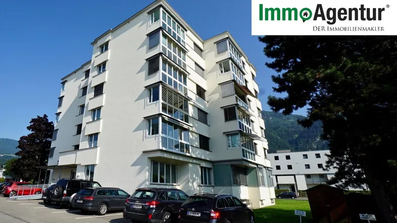 Modernes Mehrfamilienhaus mit mehreren Etagen und Parkplätzen vor einer Bergkulisse.