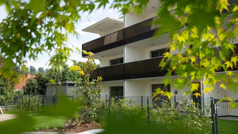 Modernes Mehrfamilienhaus mit Balkonen, umgeben von grüner Bepflanzung und Bäumen.