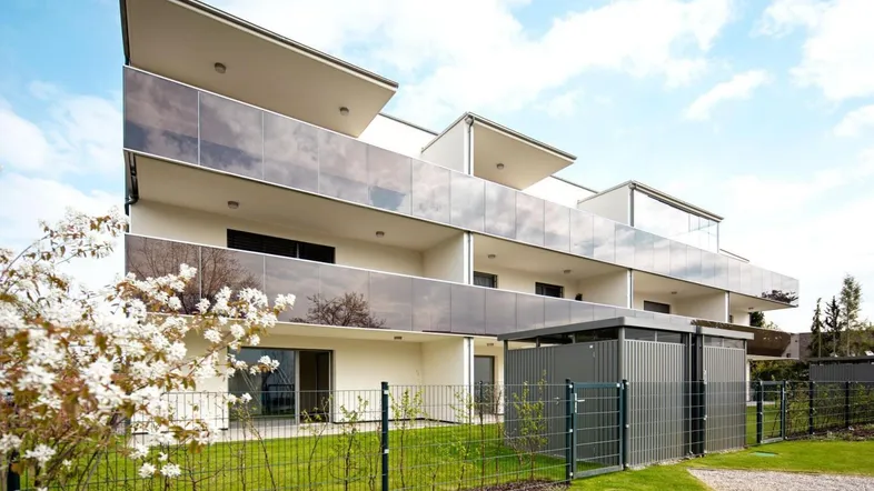 Modernes Mehrfamilienhaus mit Balkonen und Glasbrüstungen in einer grünen Umgebung.