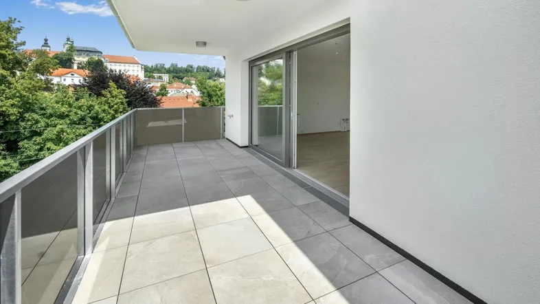 Geräumiger Balkon mit Fliesenboden, Glasgeländer und weitem Blick über die Stadt und grüne Umgebung.