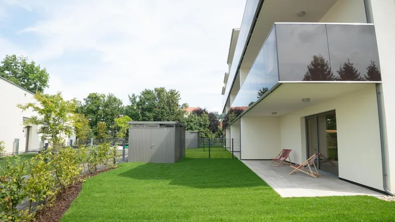 Moderne Wohnanlage mit großer Terrasse, grünem Rasen und Gartenhaus im Hintergrund.