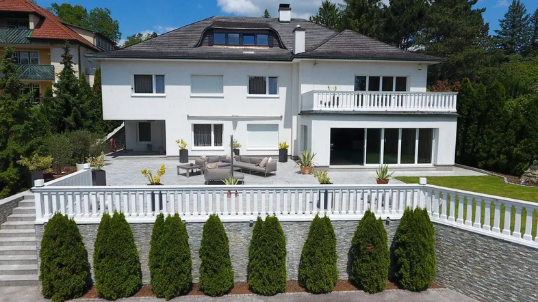 Elegante weiße Villa mit großer gepflasterter Terrasse und modernen Gartenmöbeln, umgeben von Grün.