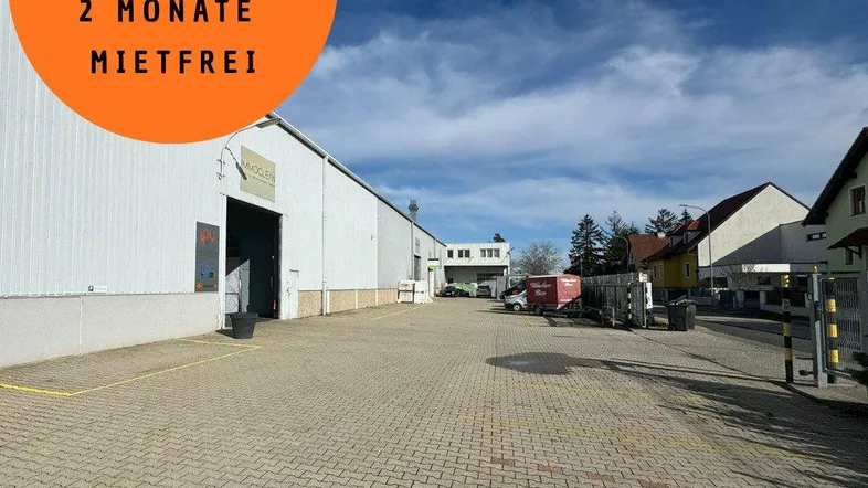 Außenansicht eines modernen Lagergebäudes mit großem gepflasterten Parkplatz unter blauem Himmel, inklusive Werbehinweis.