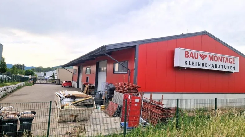 Rotes Industriegebäude mit "BAU MONTAGE" Beschriftung und umzäuntem Vorplatz mit Baumaterialien.
