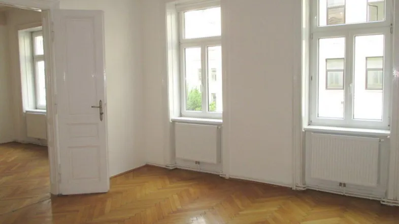 Helles Zimmer mit Fischgrätparkettboden, weißen Wänden und zwei großen Fenstern.