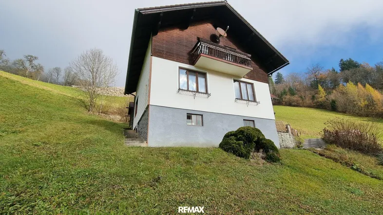 Traditionelles Haus in Hanglage mit Balkon und ländlicher Umgebung unter bewölktem Himmel.