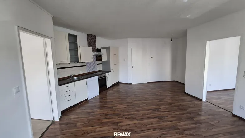 Offener Wohnbereich mit integrierter Küche, dunklem Laminatboden und weißen Schränken.