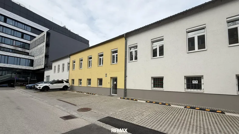 Gebäudeansicht mit gelber und weißer Fassade sowie Parkplätzen davor unter bewölktem Himmel.