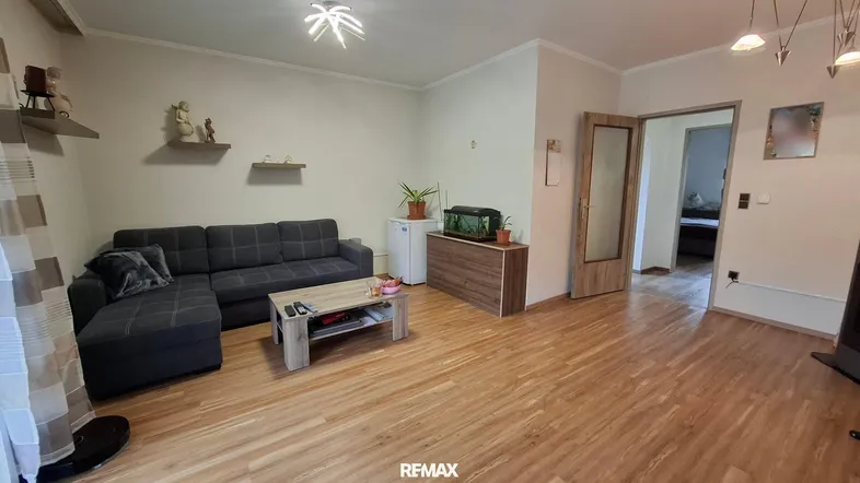 Helles Wohnzimmer mit dunklem Ecksofa, Holzboden und Zugang zu einem weiteren Raum.