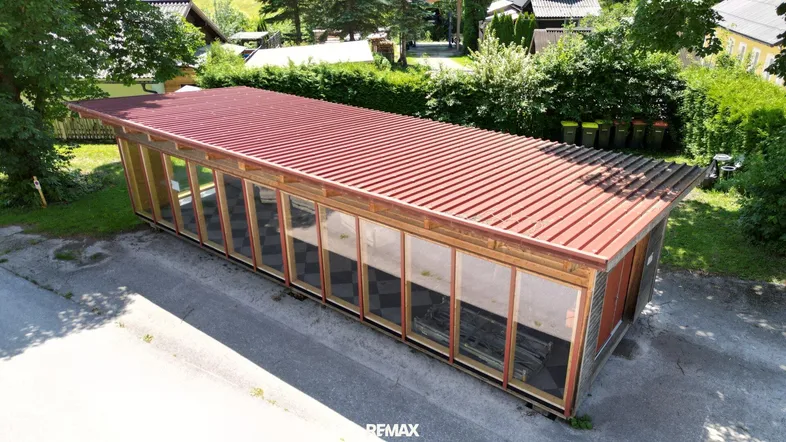 Drohnenaufnahme eines länglichen Holzgebäudes mit rotem Pultdach und großen Glasfronten.
