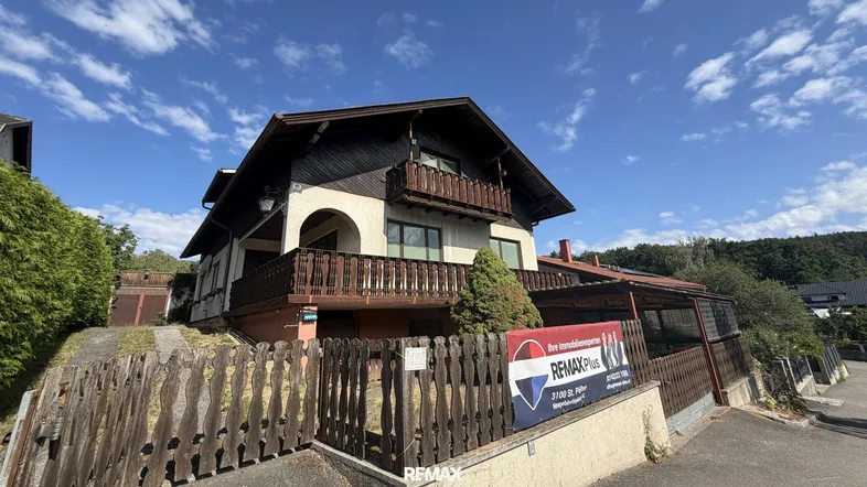 Traditionelles Haus mit Holzbalkonen und einem Maklerschild unter blauem Himmel.