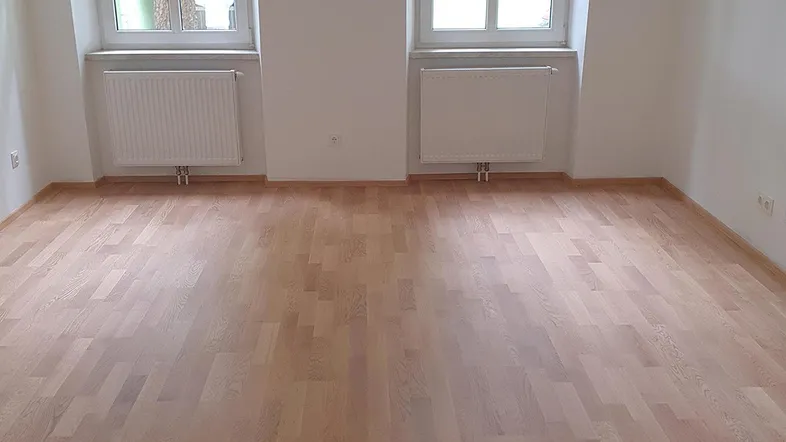 Helles Zimmer mit neuem Holzparkettboden, weißen Wänden und zwei Fenstern mit Heizkörpern.