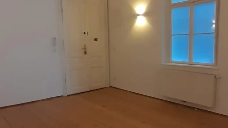 Leerer Raum mit Holzboden, weißen Wänden, einem Fenster und Wandleuchten.