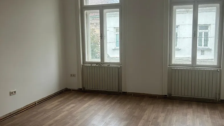 Helles Zimmer mit Holzboden und zwei großen Fenstern, bereit zur individuellen Gestaltung.
