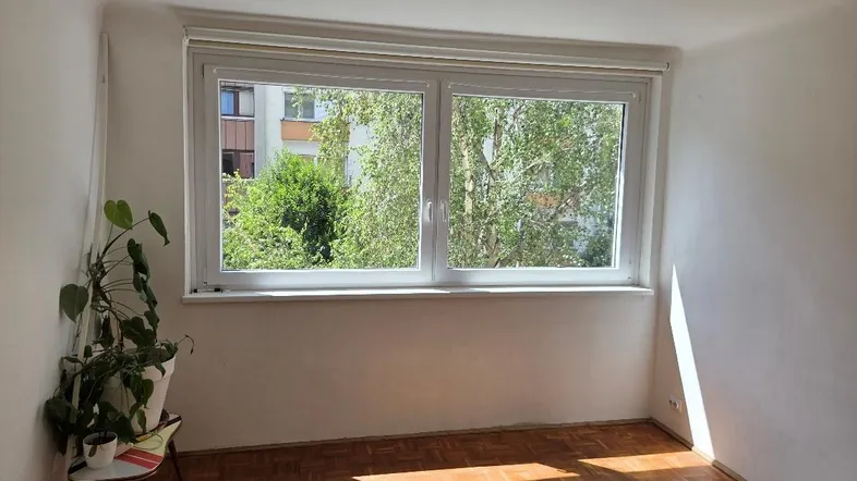 Helles Zimmer mit Parkettboden und großem Fenster, das einen Blick ins Grüne bietet.