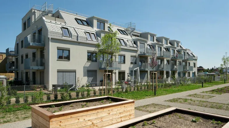 Modernes Mehrfamilienhaus mit Balkonen und Mansarddach, umgeben von gepflegten Hochbeeten und jungen Bäumen unter blauem Himmel.