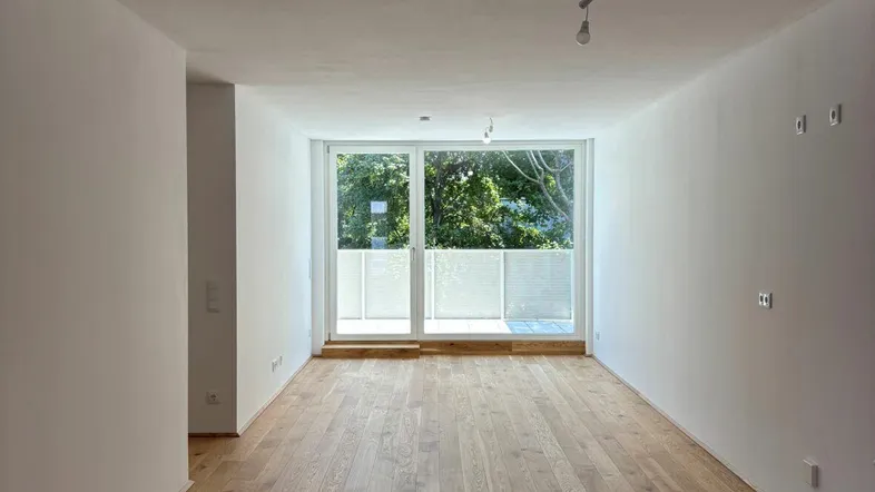 Helles, leeres Zimmer mit Holzboden und Zugang zu einem Balkon mit Blick ins Grüne.