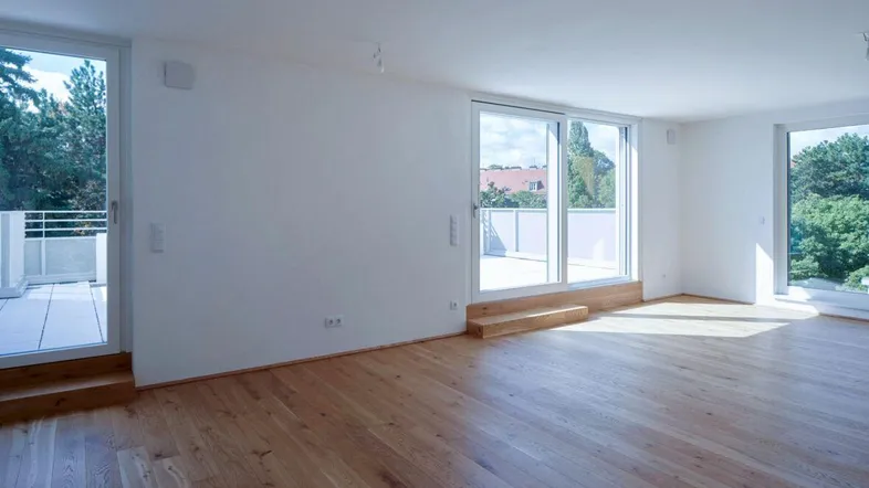 Helles, leeres Wohnzimmer mit Holzboden und großen Fenstern, die viel natürliches Licht hereinlassen.