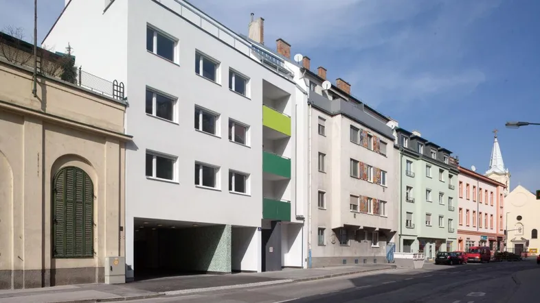 Moderne Wohngebäude an einer Straße mit angrenzenden älteren Häusern und einem Kirchturm im Hintergrund.