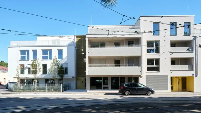 Modernes Wohngebäude mit weißer Fassade, großen Fenstern und Balkonen an einer städtischen Straße.