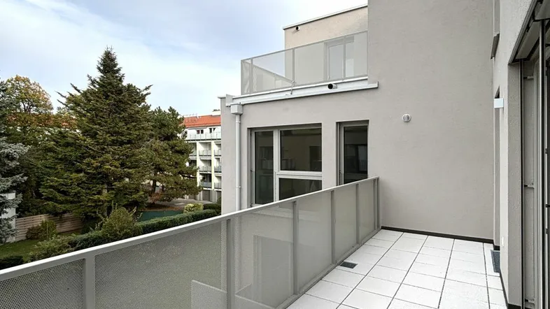 Geräumiger Balkon mit modernen Fliesen und Glasgeländer, umgeben von Grünflächen.