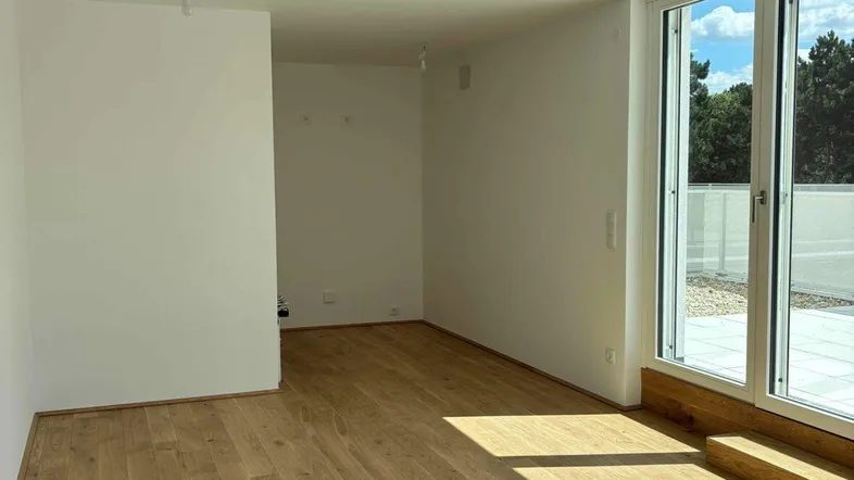 Helles Zimmer mit Holzboden und großen Fenstern, die auf eine geflieste Terrasse führen.