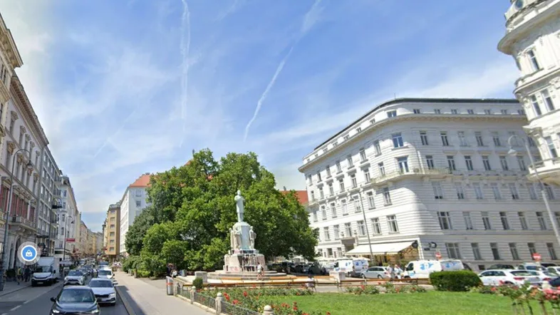 Stadtansicht mit historischen Gebäuden, einem zentralen Grünstreifen und einem Denkmal unter blauem Himmel.