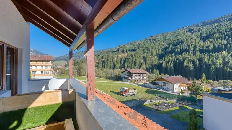 Balkon mit Holzdach und weitem Blick auf grüne Berge, Wald und umliegende Häuser.