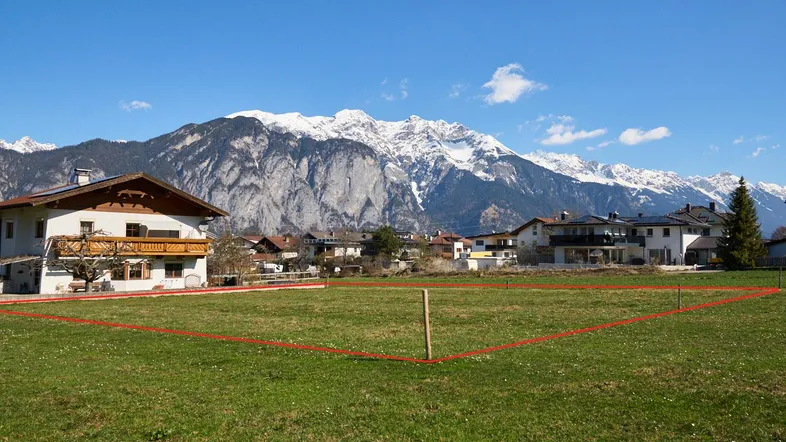 Großes grünes Grundstück mit roter Umrandung, umgeben von Häusern und majestätischen Bergen im Hintergrund.