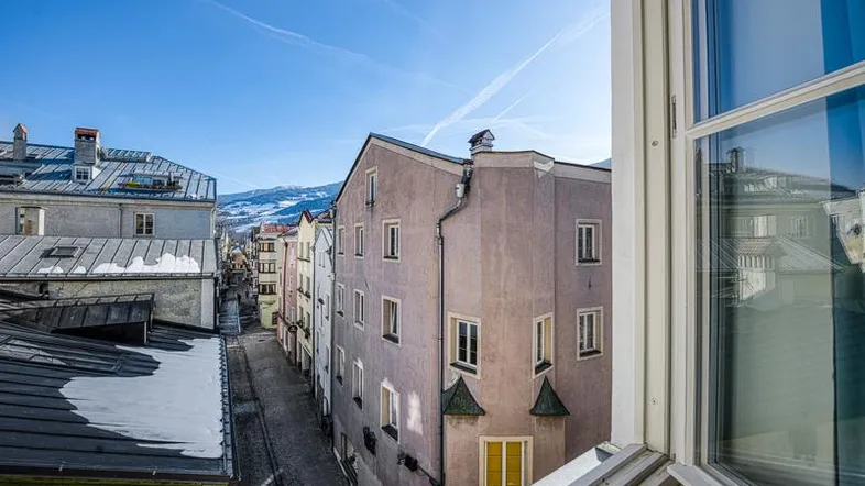 Blick aus einem Fenster auf eine historische Gasse mit Gebäuden und schneebedeckten Bergen im Hintergrund.