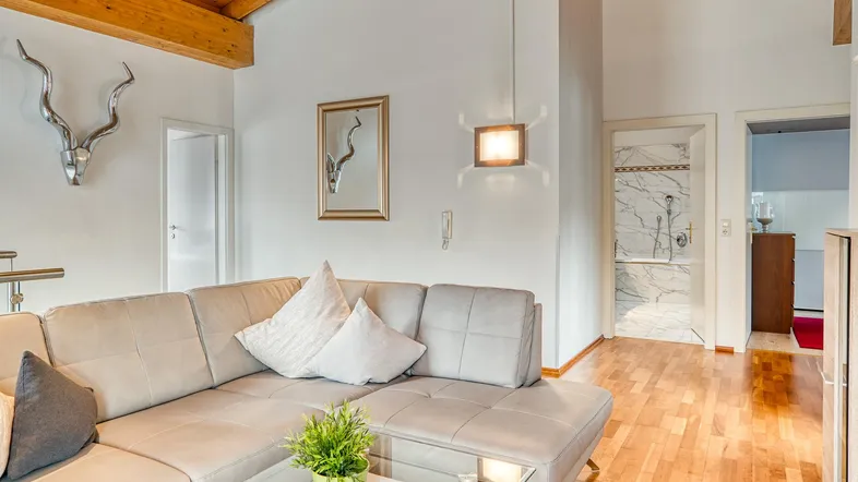 Helles Wohnzimmer mit beigem Sofa, Holzbalkendecke und Parkettboden, mit Blick auf ein Badezimmer.